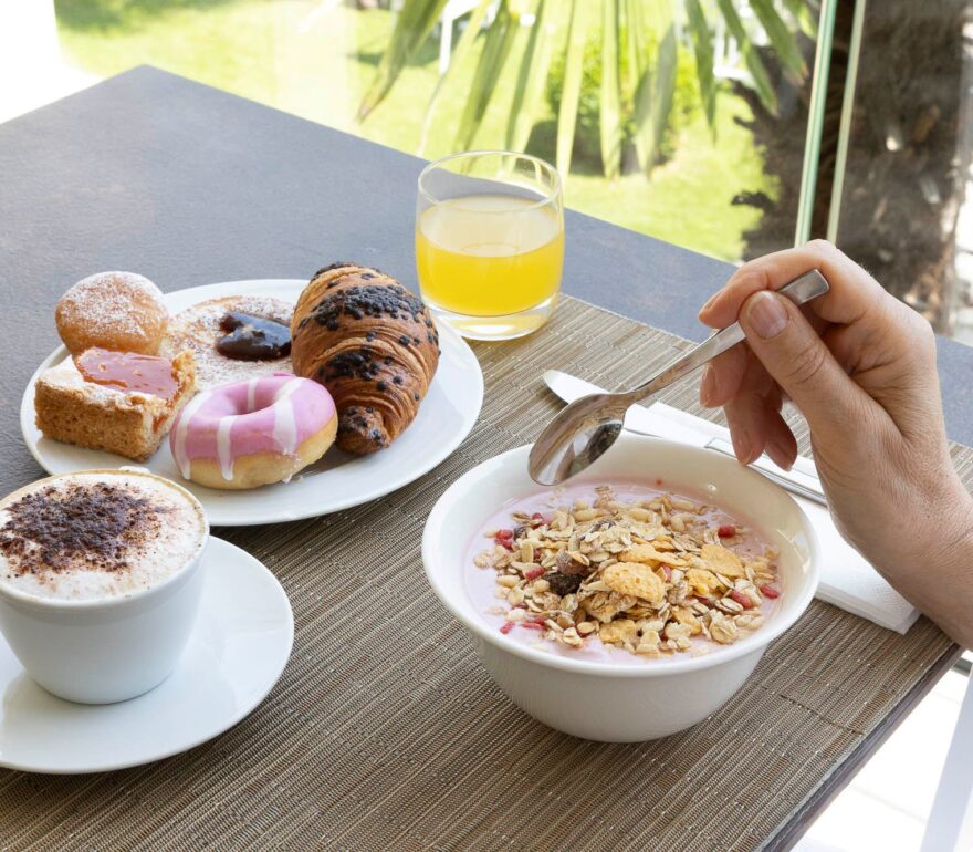 A woman in a bathrobe is having breakfast outdoors at a brown table; there are various pastries, a cup of cappuccino, and a glass of orange juice. In the background, a garden with palm trees is visible.